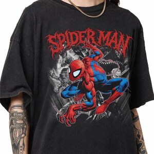 May include: A black short-sleeve t-shirt featuring a graphic print of the character Spider-Man in a crouching pose. Above the character, the word SPIDER-MAN is printed in a stylized red font. The background graphic includes black and white imagery.