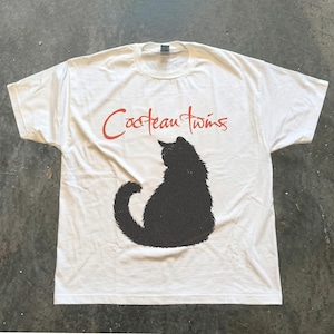 May include: A white short-sleeve t-shirt featuring a black silhouette of a fluffy cat sitting in the center. Above the cat, the text Cocteau Twins is printed in a red, cursive-style font. The shirt has a crew neckline and a standard fit.