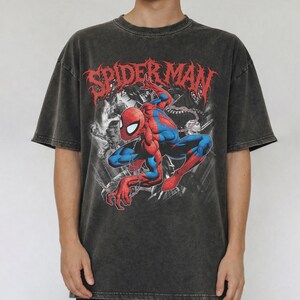 May include: A charcoal grey acid-wash t-shirt featuring a graphic print of the Marvel character Spider-Man in a crouching pose. The red text SPIDER-MAN is printed in a stylized font above the character, with black and white background imagery.
