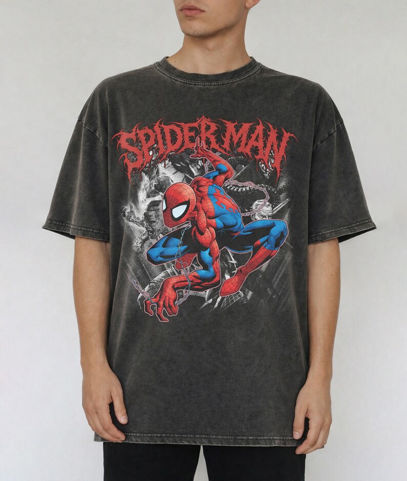 May include: A charcoal grey acid-wash t-shirt featuring a graphic print of the Marvel character Spider-Man in a crouching pose. The red text SPIDER-MAN is printed in a stylized font above the character, with black and white background imagery.
