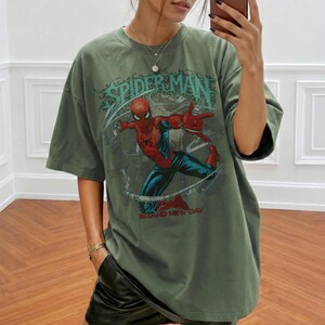 May include: An olive green short-sleeve t-shirt featuring a graphic print of the comic book character Spider-Man in a dynamic pose. The design includes the text "SPIDER-MAN" in a stylized font above the character and "BRAND NEW DAY" below.