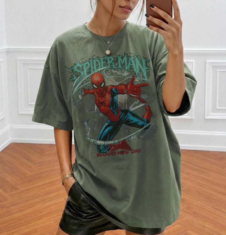 May include: An olive green short-sleeve t-shirt featuring a graphic print of the comic book character Spider-Man in a dynamic pose. The design includes the text "SPIDER-MAN" in a stylized font above the character and "BRAND NEW DAY" below.