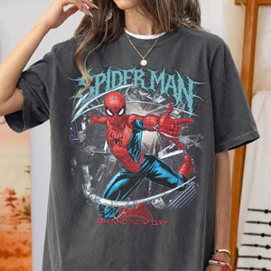 May include: A charcoal gray short-sleeve t-shirt featuring a graphic of the superhero Spider-Man in a dynamic pose. The design includes the text "SPIDER-MAN" in a stylized blue font above the character and "BRAND NEW DAY" in red text below.