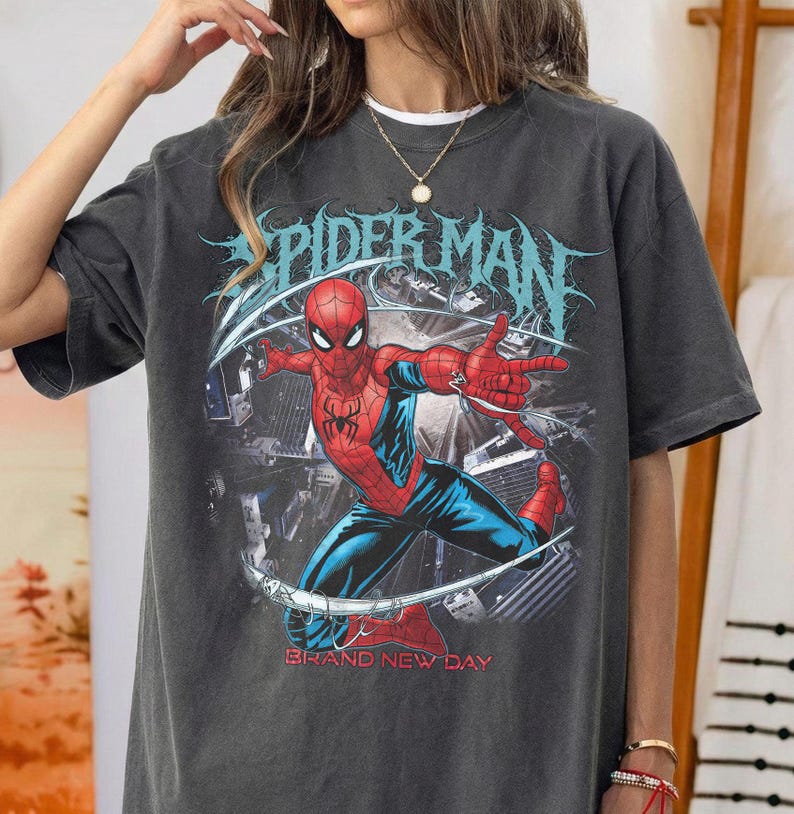 May include: A charcoal gray short-sleeve t-shirt featuring a graphic of the superhero Spider-Man in a dynamic pose. The design includes the text "SPIDER-MAN" in a stylized blue font above the character and "BRAND NEW DAY" in red text below.