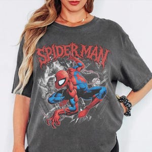 May include: A charcoal gray short-sleeve t-shirt featuring a large graphic print of the Marvel character Spider-Man in a crouching pose. The design includes the red text Spider-Man in a stylized font above the character and black and white imagery.