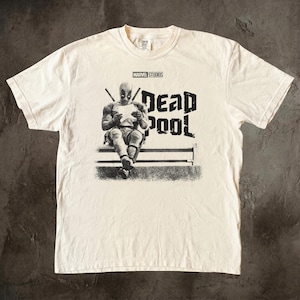 May include: A cream-colored short-sleeve t-shirt featuring a black and white graphic of Deadpool sitting on a bench reading a letter. The text MARVEL STUDIOS is printed above the character, and the word DEADPOOL is printed in large letters to the right.
