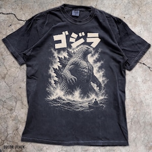 May include: A black short-sleeve t-shirt featuring a large off-white graphic print of Godzilla emerging from the ocean waves. Above the creature, the Japanese text ゴジラ is printed in a bold, distressed font. A small boat with a figure is visible.