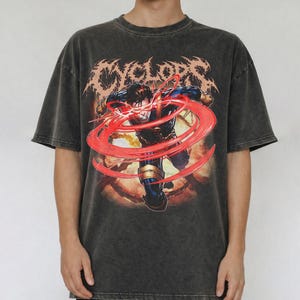 May include: A charcoal grey acid-wash t-shirt featuring a graphic print of the comic book character Cyclops. The character is shown in a dynamic pose with red energy beams circling their visor. The word CYCLOPS is printed in a jagged font above.
