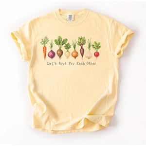 Peut inclure: Un t-shirt à manches courtes jaune pâle orné d'une illustration graphique de divers légumes racines, dont une carotte, des oignons et des radis. Sous les légumes, le texte Let's Root For Each Other est inscrit dans une police noire simple.