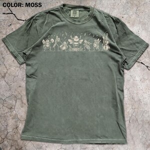 Op de afbeelding: Een mosgroen t-shirt met korte mouwen en een crèmekleurige grafische print van een groep insecten die muziekinstrumenten bespelen. Het ontwerp bevat een bij, krekels en andere insecten met muzieknoten erboven. Het shirt heeft een ronde hals.