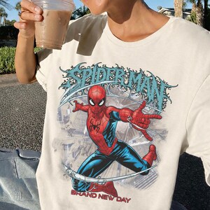 May include: A cream-colored t-shirt featuring a graphic of the superhero Spider-Man in a dynamic pose. The design includes the text SPIDER-MAN in a stylized font at the top and the words BRAND NEW DAY in red lettering at the bottom of the print.