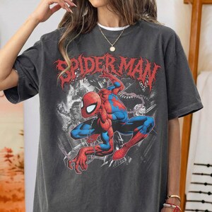 May include: A charcoal gray short-sleeve t-shirt featuring a red and blue Spider-Man graphic in a crouching pose. The text "SPIDER-MAN" is printed in a distressed red font above the character, set against a black and white background illustration.