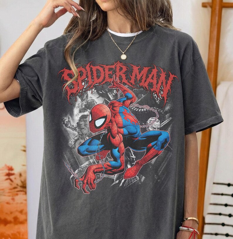 May include: A charcoal gray short-sleeve t-shirt featuring a red and blue Spider-Man graphic in a crouching pose. The text "SPIDER-MAN" is printed in a distressed red font above the character, set against a black and white background illustration.