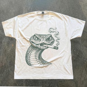 Peut inclure: Un t-shirt blanc à manches courtes orné d'une illustration graphique vert foncé représentant un serpent avec une cigarette à la bouche. Le serpent présente des écailles détaillées et des yeux perçants, avec un filet de fumée au-dessus.