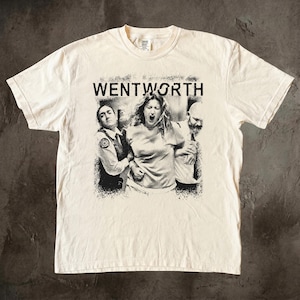 Peut inclure: Un t-shirt à manches courtes de couleur crème avec une impression graphique en noir et blanc sur le devant. Le motif représente trois personnes avec le texte WENTWORTH imprimé au-dessus dans une police grasse. Le design a un aspect usé.