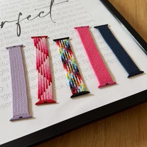 Op de afbeelding: Vijf kleurrijke horlogebandjes worden getoond tegen een ingelijste print met het woord "Perfect". De bandjes zijn onder andere lavendel, roze, een veelkleurige band, felroze en marineblauw. De achtergrond bevat songteksten.
