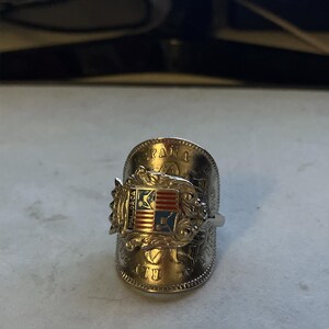 May include: A silver-toned ring with a shield design featuring red, white, and blue accents. The ring has intricate detailing and appears to be crafted from a coin. The ring is a unique piece of jewelry.