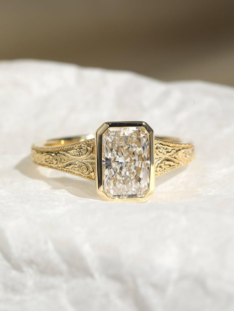 Pu&ograve; includere: Un anello in oro con una grande pietra preziosa trasparente rettangolare sfaccettata incastonata in una lunetta liscia. La fascia &egrave; realizzata in oro giallo e presenta intricati motivi a volute in rilievo sui lati, partendo dal centro.
