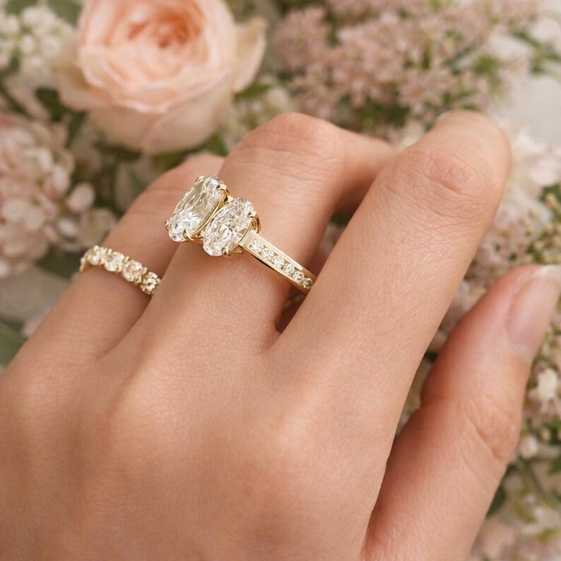 Pu&ograve; includere: Un anello di fidanzamento in oro con due grandi diamanti taglio ovale e diamanti pi&ugrave; piccoli incastonati lungo la fascia. &Egrave; visibile anche un secondo anello con diamanti rotondi pi&ugrave; piccoli. Gli anelli sono su uno sfondo floreale morbido.