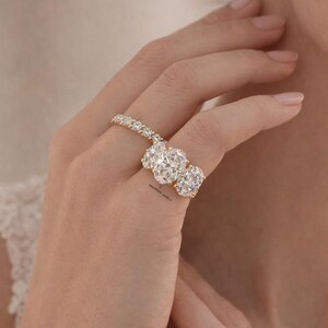 Pu&ograve; includere: Un anello d'oro con tre grandi diamanti taglio ovale e una fascia tempestata di diamanti. L'anello &egrave; indossato su un dito, con un orecchino a lobo di diamanti abbinato. Un gioiello.