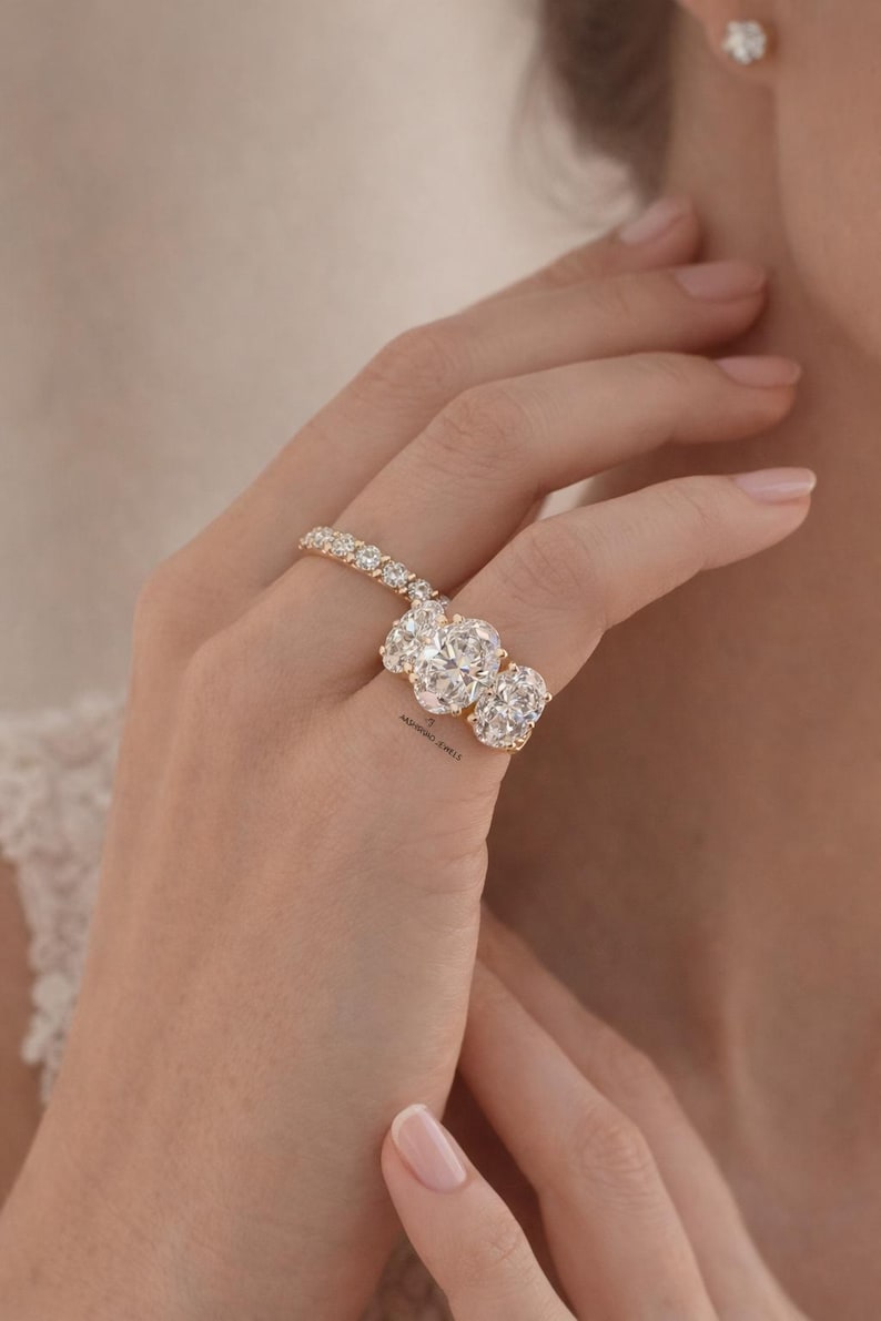 Pu&ograve; includere: Un anello d'oro con tre grandi diamanti taglio ovale e una fascia tempestata di diamanti. L'anello &egrave; indossato su un dito, con un orecchino a lobo di diamanti abbinato. Un gioiello.