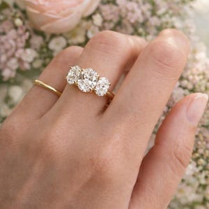 Pu&ograve; includere: Un anello d'oro con tre diamanti taglio ovale &egrave; mostrato su una mano. L'anello &egrave; ambientato su uno sfondo di fiori rosa e bianchi, creando un'estetica delicata ed elegante.