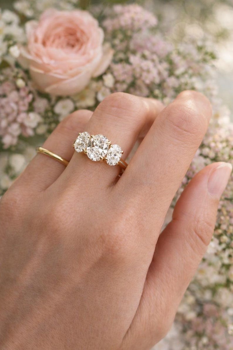 Pu&ograve; includere: Un anello d'oro con tre diamanti taglio ovale &egrave; mostrato su una mano. L'anello &egrave; ambientato su uno sfondo di fiori rosa e bianchi, creando un'estetica delicata ed elegante.