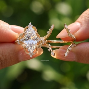 Puede incluir: Un conjunto de anillos de oro con una gran piedra central de corte marquesa rodeada por un marco de filigrana decorativo con pequeños diamantes engastados en pavé. La banda incluye extensiones en forma de rama adornadas con diamantes diminutos. El texto AASHIRVAD JEWELS es visible.
