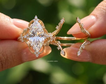 May include: A marquise-cut diamond engagement ring with a gold band and detailed filigree. Two curved diamond-studded bands accompany the ring. The rings are displayed against a blurred green backdrop.