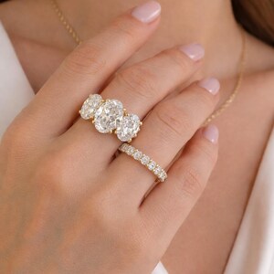 Pu&ograve; includere: Una mano mostra due anelli d'oro. Uno presenta tre diamanti taglio ovale, l'altro &egrave; una fascia di diamanti pi&ugrave; piccoli. &Egrave; visibile anche una collana d'oro.
