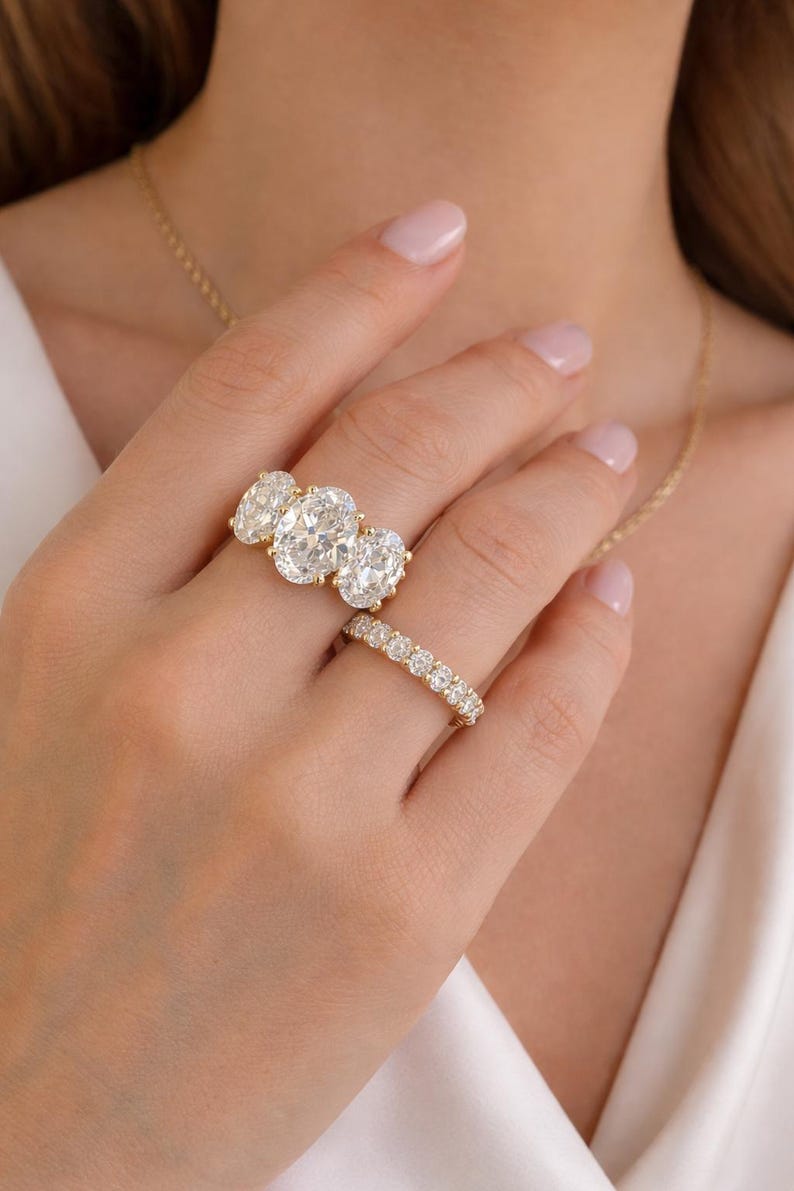 Pu&ograve; includere: Una mano mostra due anelli d'oro. Uno presenta tre diamanti taglio ovale, l'altro &egrave; una fascia di diamanti pi&ugrave; piccoli. &Egrave; visibile anche una collana d'oro.