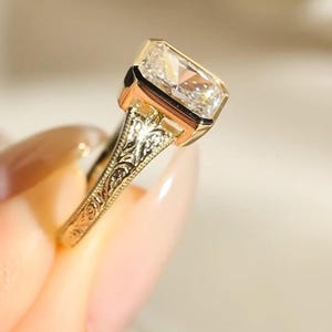 Pu&ograve; includere: Un anello in oro con una pietra preziosa trasparente a taglio radiante rettangolare in una montatura a castone. La fascia &egrave; realizzata in oro giallo e presenta intricati motivi a volute floreali incisi sui lati del gambo.