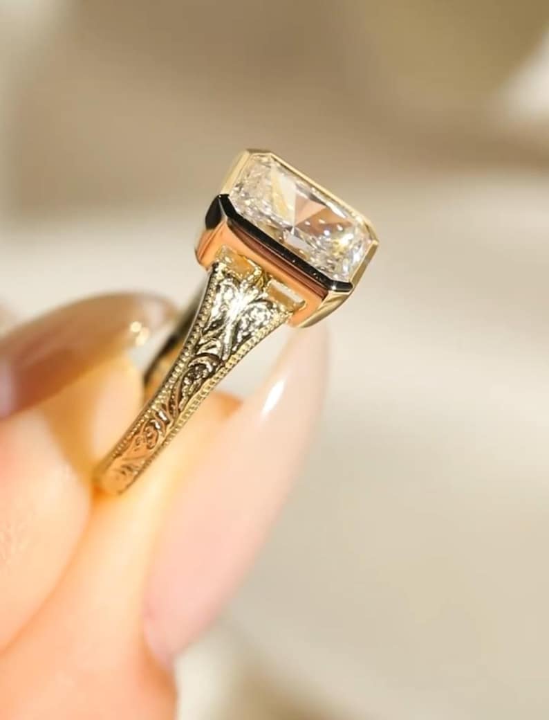 Pu&ograve; includere: Un anello in oro con una pietra preziosa trasparente a taglio radiante rettangolare in una montatura a castone. La fascia &egrave; realizzata in oro giallo e presenta intricati motivi a volute floreali incisi sui lati del gambo.