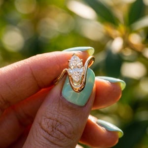 May include: A gold ring featuring two pear-shaped diamonds. The ring's band has a unique, flowing design. The diamonds sparkle against a blurred green background, suggesting an outdoor setting. The ring is held up by a person's hand.