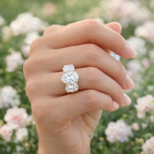 Pu&ograve; includere: Un anello d'oro con tre diamanti taglio ovale. I diamanti sono disposti in fila, con il diamante pi&ugrave; grande al centro. L'anello &egrave; su un dito, con uno sfondo sfocato di fiori.
