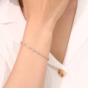 Puede incluir: Una delicada pulsera de cadena dorada con una serie de pequeñas piedras redondas y transparentes engastadas a intervalos regulares a lo largo de la banda. La pulsera se asegura con un cierre y presenta un diseño lineal y sencillo.