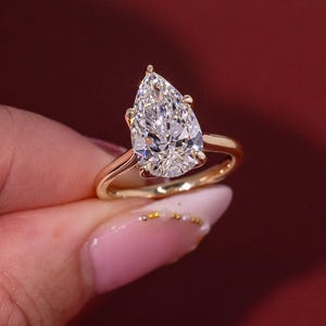 May include: A gold ring featuring a large, pear-shaped diamond. The diamond is clear and faceted, reflecting light. The ring has a simple, elegant design with a gold band. The ring is held against a red background.