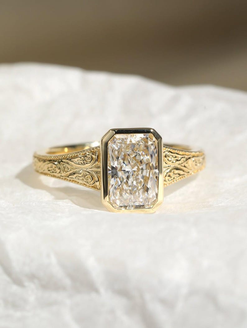 Pu&ograve; includere: Un anello in oro con una grande pietra preziosa trasparente rettangolare sfaccettata incastonata in una lunetta liscia. La fascia &egrave; realizzata in oro giallo e presenta intricati motivi a volute in rilievo sui lati, partendo dal centro.