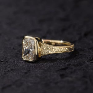 Pu&ograve; includere: Un anello in oro con una grande pietra preziosa trasparente dal taglio radiante rettangolare, montata in una lunetta liscia. La fascia &egrave; realizzata in oro giallo e presenta intricati motivi a volute incisi sui lati del gambo.