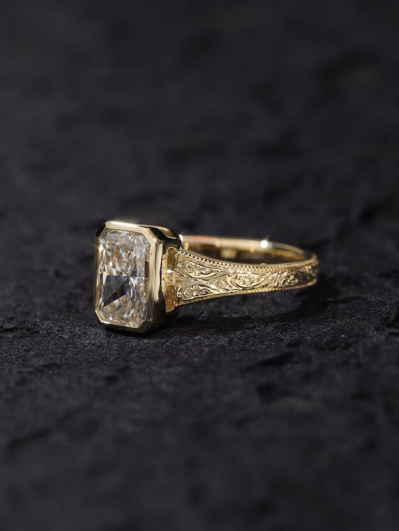 Pu&ograve; includere: Un anello in oro con una grande pietra preziosa trasparente dal taglio radiante rettangolare, montata in una lunetta liscia. La fascia &egrave; realizzata in oro giallo e presenta intricati motivi a volute incisi sui lati del gambo.