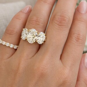Pu&ograve; includere: Primo piano di una mano che indossa due anelli d'oro. L'anello principale presenta tre diamanti taglio ovale incastonati in una fascia d'oro. Un secondo anello, anch'esso in oro, &egrave; incastonato con diamanti rotondi pi&ugrave; piccoli. La mano ha unghie curate.