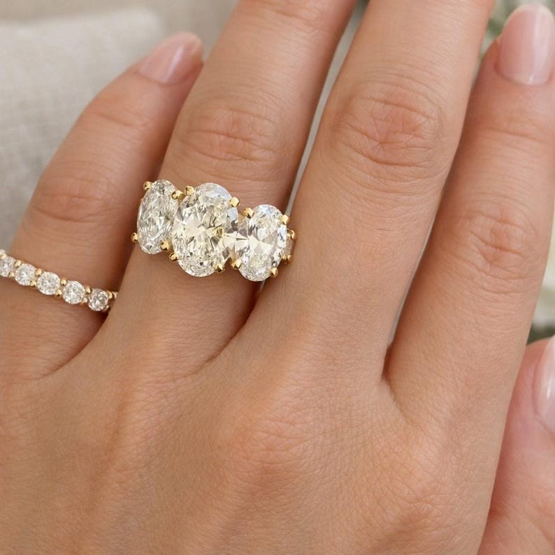 Pu&ograve; includere: Primo piano di una mano che indossa due anelli d'oro. L'anello principale presenta tre diamanti taglio ovale incastonati in una fascia d'oro. Un secondo anello, anch'esso in oro, &egrave; incastonato con diamanti rotondi pi&ugrave; piccoli. La mano ha unghie curate.