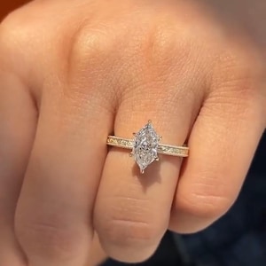 May include: A marquise-cut diamond engagement ring. The diamond is set in a gold band with a delicate, patterned design. The ring is shown on a person's finger, highlighting the intricate details of the jewelry.