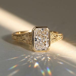 Pu&ograve; includere: Un anello in oro con una pietra centrale rettangolare taglio radiante incastonata in una lunetta liscia. La fascia in oro giallo presenta intricati motivi a volute incisi sui lati, che catturano la luce creando un effetto prismatico.