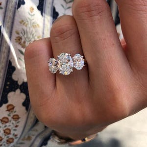 Pu&ograve; includere: Un anello di fidanzamento a tre pietre con un grande diamante ovale affiancato da due diamanti rotondi pi&ugrave; piccoli. L'anello &egrave; incastonato in una fascia d'argento, che mette in risalto la brillantezza dei diamanti. L'anello &egrave; tenuto da una mano.