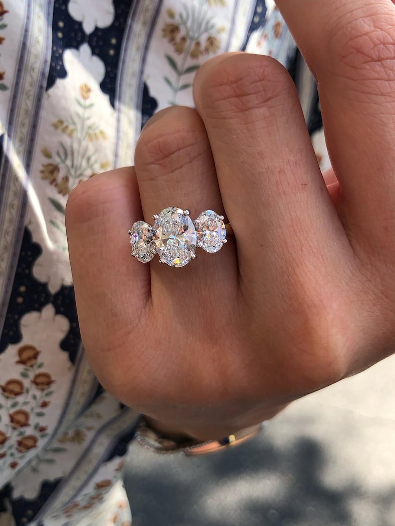 Pu&ograve; includere: Un anello di fidanzamento a tre pietre con un grande diamante ovale affiancato da due diamanti rotondi pi&ugrave; piccoli. L'anello &egrave; incastonato in una fascia d'argento, che mette in risalto la brillantezza dei diamanti. L'anello &egrave; tenuto da una mano.