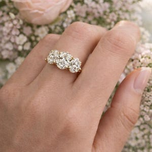 Pu&ograve; includere: Un anello d'oro con tre grandi diamanti scintillanti. L'anello &egrave; indossato su una mano, con uno sfondo floreale di fiori rosa e bianchi. I diamanti sono di forma ovale e sono posizionati vicini.