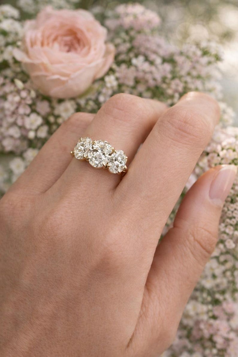 Pu&ograve; includere: Un anello d'oro con tre grandi diamanti scintillanti. L'anello &egrave; indossato su una mano, con uno sfondo floreale di fiori rosa e bianchi. I diamanti sono di forma ovale e sono posizionati vicini.