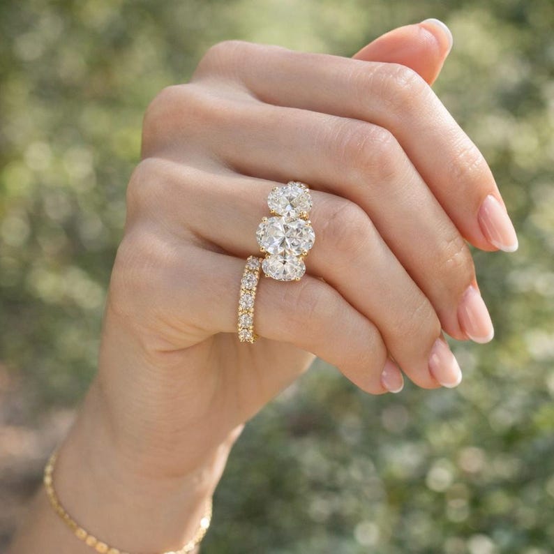 Pu&ograve; includere: Una mano mostra due anelli d'oro. Uno presenta tre diamanti taglio ovale, e l'altro &egrave; una fascia con piccoli diamanti rotondi. &Egrave; visibile anche un braccialetto d'oro.