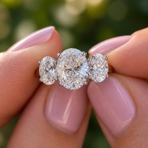 Pu&ograve; includere: Anello di fidanzamento in oro con tre diamanti taglio ovale. Il diamante centrale &egrave; pi&ugrave; grande delle due pietre laterali. L'anello &egrave; tenuto su uno sfondo verde sfocato, evidenziando la brillantezza dei diamanti.